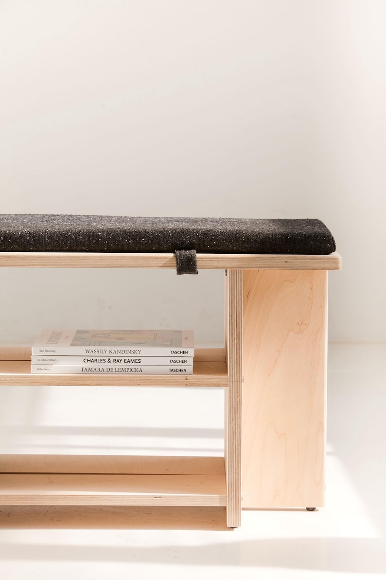 May include: A light-colored plywood bench featuring a dark gray, textured fabric cushion on top. The bench includes a lower shelf holding three books titled Wassily Kandinsky, Charles & Ray Eames, and Tamara de Lempicka, all published by Taschen.