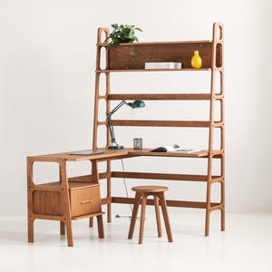 Op de afbeelding: Een L-vormig bureau van lichtbruin hout met een geïntegreerde hoge kast. Het bureau heeft een lade aan de linkerkant en een bijpassende houten kruk. De kast bevat meerdere horizontale planken met een groene plant, een gele vaas, een donkergroene bureaulamp en een open boek.