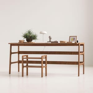 May include: A wooden desk with a rectangular top and open side frames, featuring a matching two-tiered wooden stool placed in front. The desk surface holds a potted green plant, a white lamp, a small sculpture, and a framed picture.
