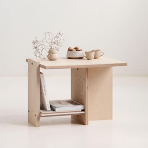Può includere: Un tavolino in legno chiaro con un ripiano, con libri. Il piano del tavolo mostra un piccolo vaso con rami secchi, una ciotola di noci e due piccole tazze. Il tavolo è largo circa 61 cm e alto 51 cm.