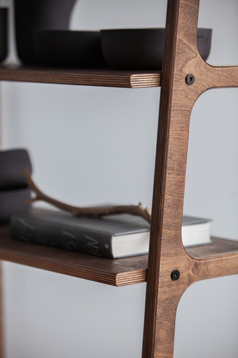 Op de afbeelding: Een houten ladderrek met vier planken. De planken zijn gemaakt van multiplex en hebben een donkerbruine vlek. Het rek is gemaakt van massief hout en heeft een natuurlijke afwerking. Het rek is leeg behalve voor een boek op de onderste plank.