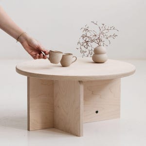 May include: A round light-coloured plywood coffee table with a cross-shaped base. On the table sits a small, smooth, beige ceramic vase containing a dried plant branch. Two small, speckled, light-brown ceramic mugs are positioned on the surface.