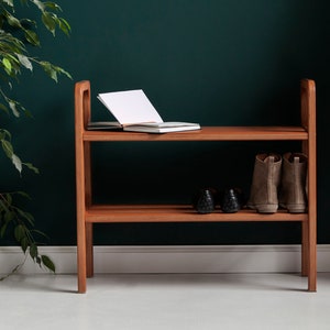 May include: A two-tiered wooden shoe rack. An open notebook rests on the top shelf. The lower shelf displays two pairs of shoes and boots. The rack is made of light brown wood and is against a dark green wall.