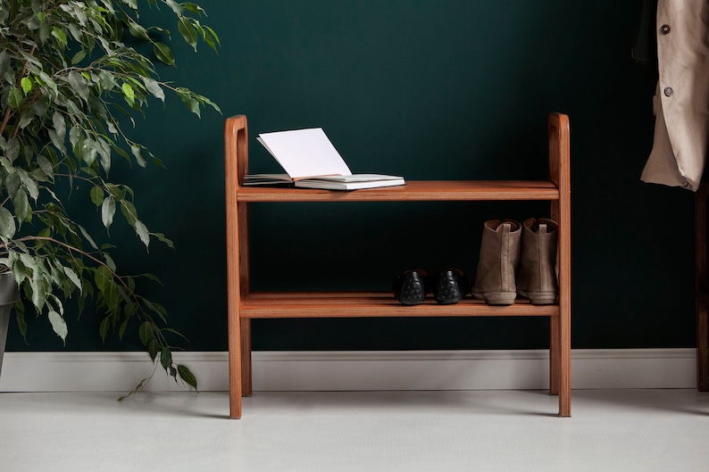 May include: A two-tiered wooden shoe rack. An open notebook rests on the top shelf. The lower shelf displays two pairs of shoes and boots. The rack is made of light brown wood and is against a dark green wall.