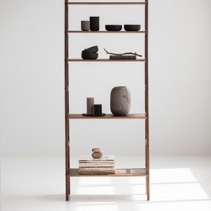 Op de afbeelding: Een houten ladderrek met vijf planken. De planken zijn gevuld met verschillende items, waaronder kaarsen, kommen, een vaas en een stapel boeken. Het rek staat tegen een witte muur.