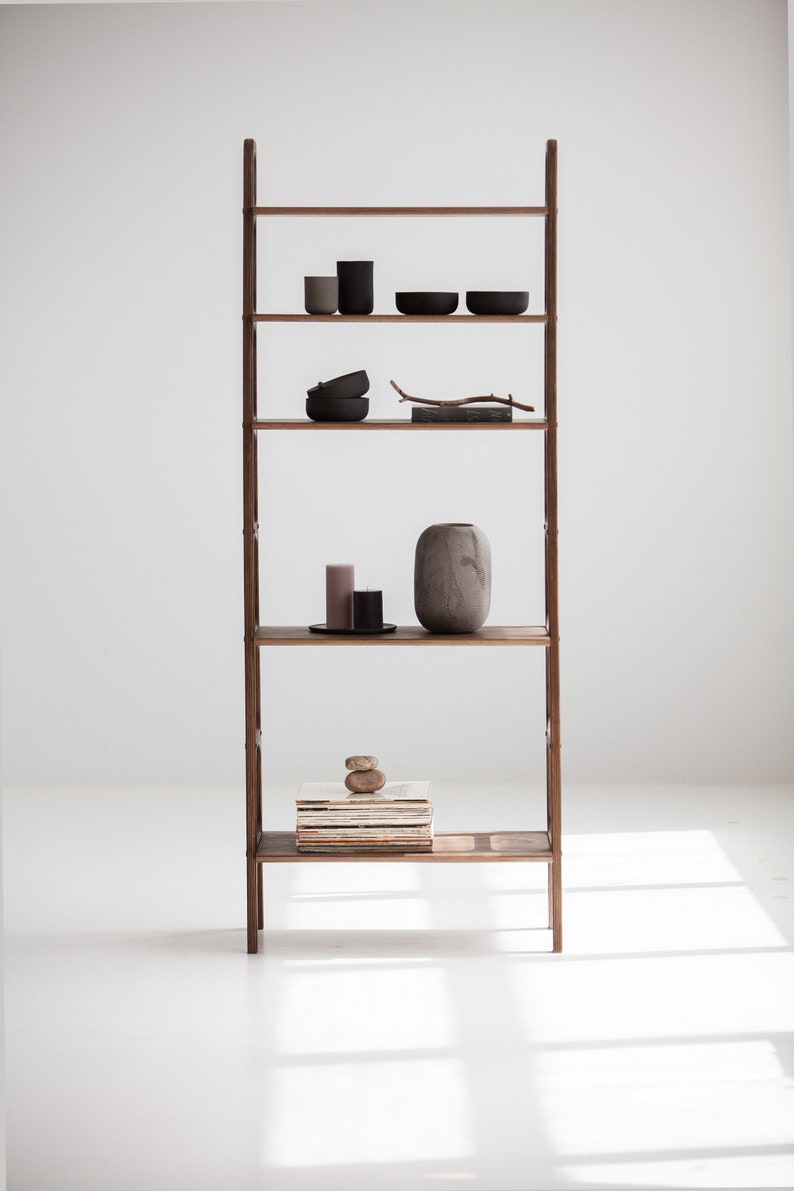 Op de afbeelding: Een houten ladderrek met vijf planken. De planken zijn gevuld met verschillende items, waaronder kaarsen, kommen, een vaas en een stapel boeken. Het rek staat tegen een witte muur.