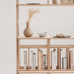 May include: A wooden bookshelf with multiple shelves, featuring a vase with dried flowers, a bowl, and a framed picture. The bottom shelf is filled with books, including titles like "The Iconic Interior", "Cimplicity", and "Oculus".