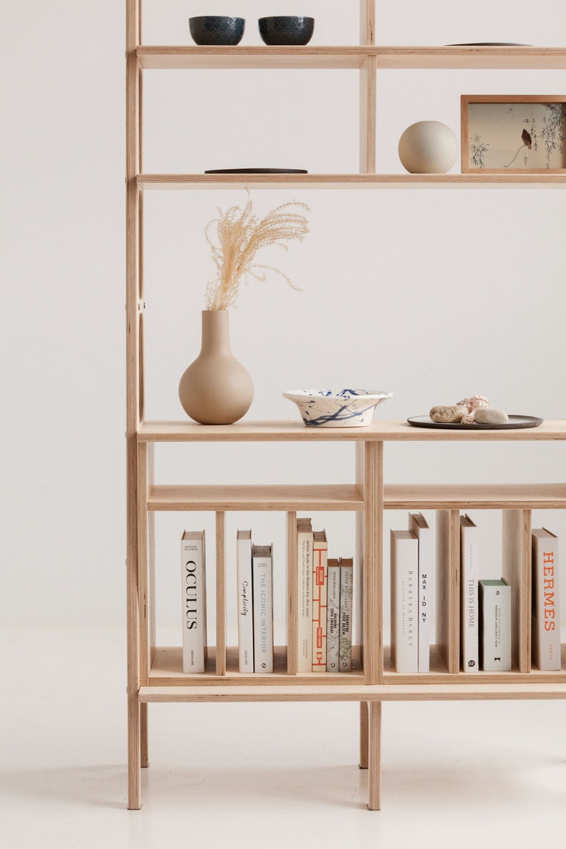 May include: A wooden bookshelf with multiple shelves, featuring a vase with dried flowers, a bowl, and a framed picture. The bottom shelf is filled with books, including titles like "The Iconic Interior", "Cimplicity", and "Oculus".