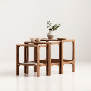 May include: A set of three nested wooden tables with a light brown finish. Each table features four legs with rounded cutouts at the top corners. The tables are arranged in a stepped formation, with a small vase and two mugs placed on top.