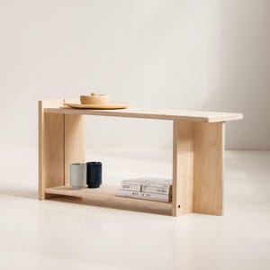 以下が含まれることがあります： 棚付きの明るい色の木製ベンチ。 ベンチは長方形の天板と2つの垂直サポートがあります。 小さな木製のボウルとプレートが上にあります。 2つの小さなカップと本の束が棚にあります。