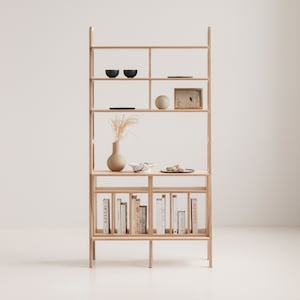 May include: A light wood bookshelf with five shelves. The top shelf has two small blue bowls and a framed picture. The second shelf has a small round white object and a framed picture. The third shelf has a vase with dried flowers and a small plate. The fourth shelf has a row of books with the titles "Herm&egrave;s" and "Studio" visible. The bookshelf is standing on a white surface.