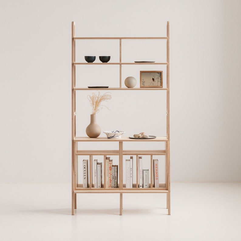 May include: A light wood bookshelf with five shelves. The top shelf has two small blue bowls and a framed picture. The second shelf has a small round white object and a framed picture. The third shelf has a vase with dried flowers and a small plate. The fourth shelf has a row of books with the titles "Herm&egrave;s" and "Studio" visible. The bookshelf is standing on a white surface.
