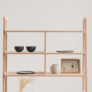 May include: A light wood shelf unit with four shelves. The top shelf has two small blue bowls and a round black plate. The second shelf has a framed print with a bird and branches. The third shelf has a round light grey vase. The bottom shelf is empty.