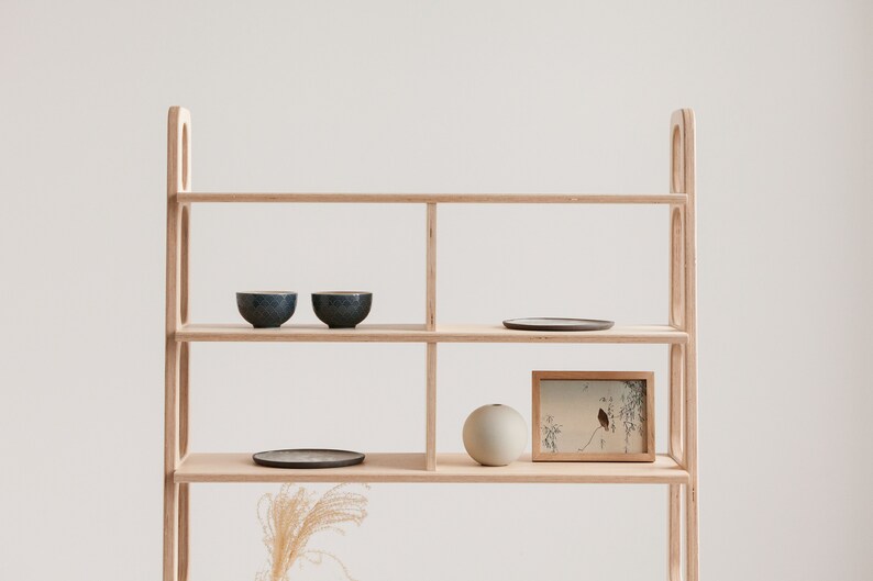 May include: A light wood shelf unit with four shelves. The top shelf has two small blue bowls and a round black plate. The second shelf has a framed print with a bird and branches. The third shelf has a round light grey vase. The bottom shelf is empty.