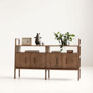 May include: A wooden sideboard featuring four cabinet doors with vertical handles, an open shelf above, and tall legs. On the top surface sit a framed picture, two small bear-shaped figurines, a green potted plant, and a small wooden bowl.