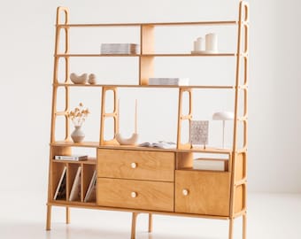 May include: A light-colored wooden bookshelf with multiple shelves, drawers, and a cabinet. The shelves display books, decorative items, and candles. The unit has a mid-century modern design with a light wood finish and rounded edges.