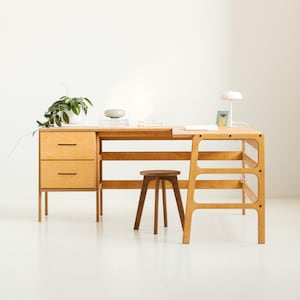 Op de afbeelding: Een licht houten bureau met een lade-eenheid en een schapeenheid. Het bureau heeft een witte lamp en een kleine kruk.