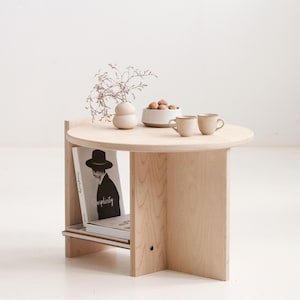 Peut inclure: Une table d'appoint en bois clair avec un plateau circulaire et une étagère inférieure. Sur la table se trouvent un petit vase beige avec des branches séchées, un bol de noix et deux tasses beiges. Un livre intitulé Simplicity est posé sur l'étagère.