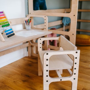 May include: A light-colored wooden chair with a flat seat and backrest, featuring a frame secured by dark screws. The chair is positioned in front of a matching wooden desk and a tall, multi-level wooden structure with open rectangular panels.