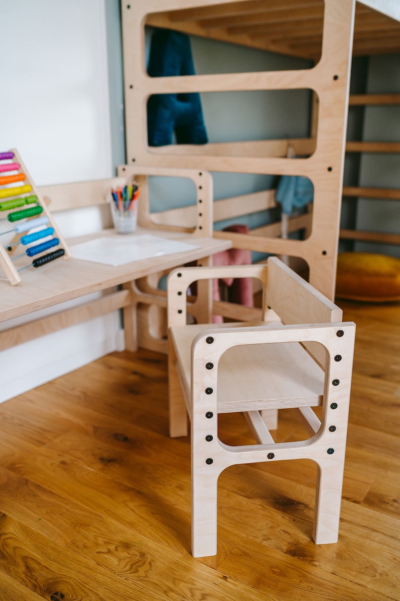 May include: A light-colored wooden chair with a flat seat and backrest, featuring a frame secured by dark screws. The chair is positioned in front of a matching wooden desk and a tall, multi-level wooden structure with open rectangular panels.