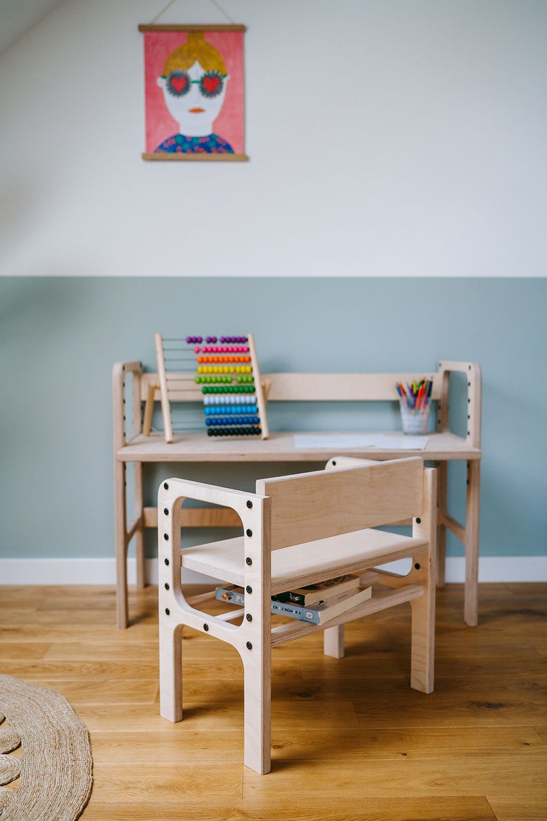May include: A light-colored wooden desk and a matching wooden chair with black hardware. The desk holds a colorful abacus and a cup of pencils. The chair features a lower shelf holding books. The furniture has a smooth, natural wood finish.