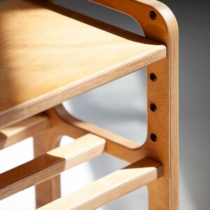 May include: A light-colored plywood stool featuring a smooth flat seat and a lower horizontal support bar. The structure is held together by visible black metal bolts integrated into the side frame, showcasing the layered wood grain edges.