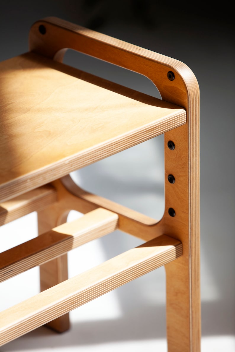 May include: A light-colored plywood stool featuring a smooth flat seat and a lower horizontal support bar. The structure is held together by visible black metal bolts integrated into the side frame, showcasing the layered wood grain edges.