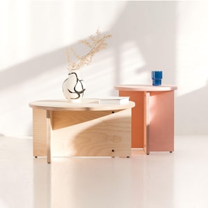 Op de afbeelding: Een lage, ronde salontafel van lichtgekleurd hout met een platte voet en een hogere, ronde bijzettafel in roze. Een witte keramische vaas met zwarte abstracte lijnen bevat gedroogde bladeren en een klein blauw glazen bekertje.