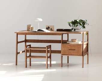 May include: A wooden desk with a drawer and a shelf underneath. The desk has a light wood finish and is made of plywood. There is a small plant on the desk, a lamp, and a book.