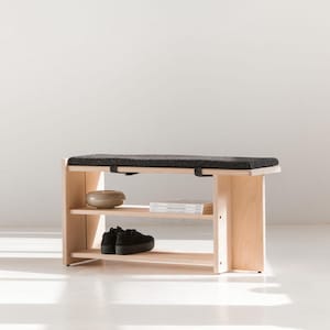 Può includere: Una panca in legno chiaro con un cuscino rettangolare dalla texture nera. La panca presenta due ripiani aperti sottostanti, sui quali sono posizionati una piccola ciotola rotonda in ceramica, una pila di libri e un paio di scarpe nere.