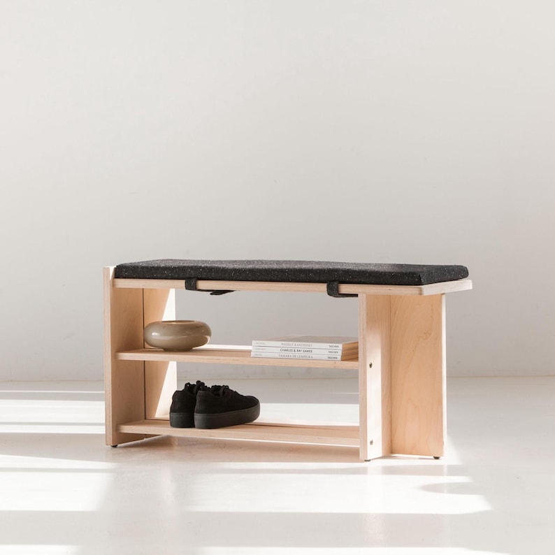 May include: A light-colored wooden bench featuring a rectangular black textured seat cushion. The bench includes two open shelves underneath, with a small round ceramic bowl, a stack of books, and a pair of black platform sneakers placed on them.