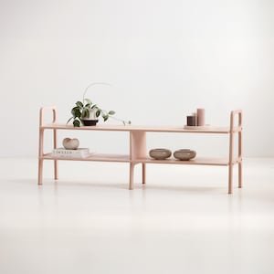 Peut inclure: Étagère en bois rose clair à deux niveaux avec un support central. L'étagère supérieure contient une plante en pot, des bougies et des bols décoratifs. L'étagère inférieure présente des livres et des bols. Le meuble a un design minimaliste.