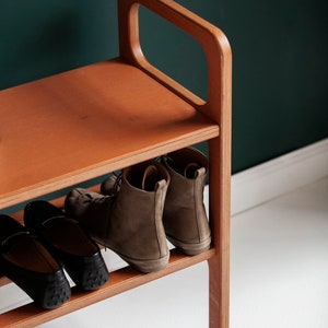 May include: A wooden shoe rack with a handle, holding several pairs of shoes. The rack is made of light brown wood and has three shelves. The shoes include black loafers and brown ankle boots. The background is a dark teal wall.