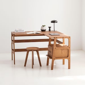 Op de afbeelding: Een licht houten hoekbureau met een plank en een lade. Op het bureau staan een laptop, boeken en een kleine plant. Voor het bureau staat een kleine houten kruk.