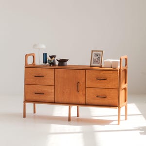 May include: A light brown wooden sideboard featuring four drawers with thin black horizontal handles, a central cabinet door with a vertical black handle, and integrated side frame supports. The top surface holds a white lamp, small bowls, a framed picture, and white boxes.