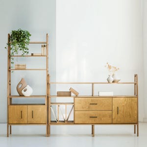 May include: A light wood shelving unit with a tall vertical section featuring open shelves and two lower cabinet doors, and a wider horizontal section with open shelving, two drawers, and a cabinet door, all accented with thin black metal handles.