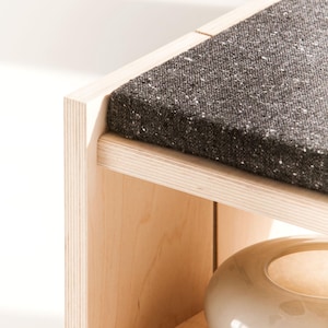 May include: A light-colored wooden bench featuring a dark charcoal gray textured fabric cushion on the top surface. The bench has a lower shelf that holds a smooth, round, off-white ceramic bowl with a glossy finish and a wide opening.