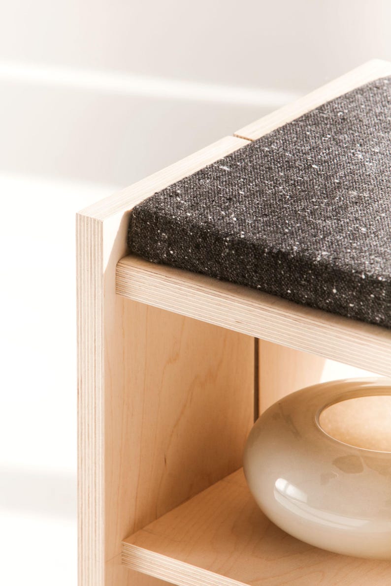 May include: A light-colored wooden bench featuring a dark charcoal gray textured fabric cushion on the top surface. The bench has a lower shelf that holds a smooth, round, off-white ceramic bowl with a glossy finish and a wide opening.