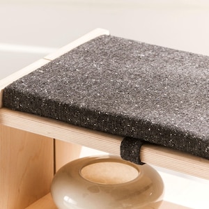 May include: A light-colored wooden bench featuring a rectangular seat cushion upholstered in a dark charcoal gray fabric with subtle white flecks. The cushion is secured to the wooden frame with a matching dark gray fabric strap.