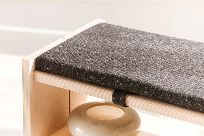 May include: A light-colored wooden bench featuring a rectangular seat cushion upholstered in a dark charcoal gray fabric with subtle white flecks. The cushion is secured to the wooden frame with a matching dark gray fabric strap.