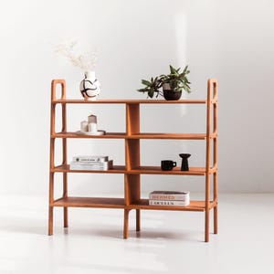 Puede incluir: Una estantería de madera con cinco estantes, que incluye un jarrón blanco con marcas negras, una planta en maceta, velas, libros y dos tazas de cerámica negras. La estantería está hecha de madera marrón claro y tiene un diseño moderno.