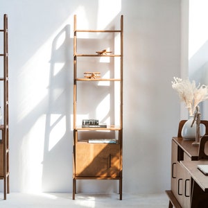 Può includere: Due scaffali in legno a forma di scala con più ripiani e una porta di un armadio nella parte inferiore. Gli scaffali sono vuoti, ad eccezione di alcuni libri e piccoli oggetti decorativi. Gli scaffali sono contro un muro bianco.