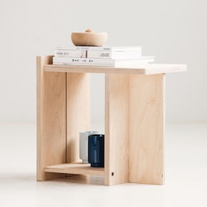 May include: A light-colored wooden side table with a shelf and a rectangular top. The table holds books, a small wooden bowl, and two ceramic mugs. The table has a modern, minimalist design.