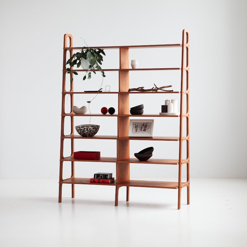 Wide mid century modern wooden bookcase with seven shelves, styled with books, ceramics and a plant, handmade from Finnish birch plywood in a warm wood finish, against a white wall.