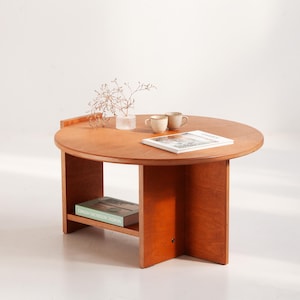 Op de afbeelding: Een ronde houten salontafel met een warme, roodbruine afwerking. De tafel heeft een onderste plank en een kleine, verhoogde rand. Twee kleine mokken, een tijdschrift en een vaas met decoratieve takken staan op het tafelblad. De tafel heeft een diameter van ongeveer 76 cm.