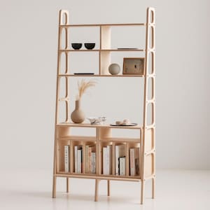 May include: A tall, light wood bookshelf with open shelves and a lower cabinet with doors. The shelves are filled with books, decorative bowls, and a framed picture. The cabinet has a light wood finish and is supported by four legs.