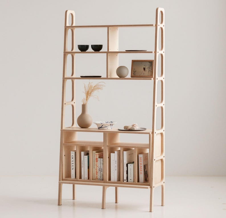 May include: A tall, light wood bookshelf with open shelves and a lower cabinet with doors. The shelves are filled with books, decorative bowls, and a framed picture. The cabinet has a light wood finish and is supported by four legs.