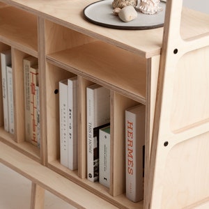 May include: A light wood bookshelf with a slanted back and a lower shelf. The bookshelf has several compartments and is filled with books, including "This is Home", "Max ID NY", "Barber & Osgerby", and "Hermes Paris".