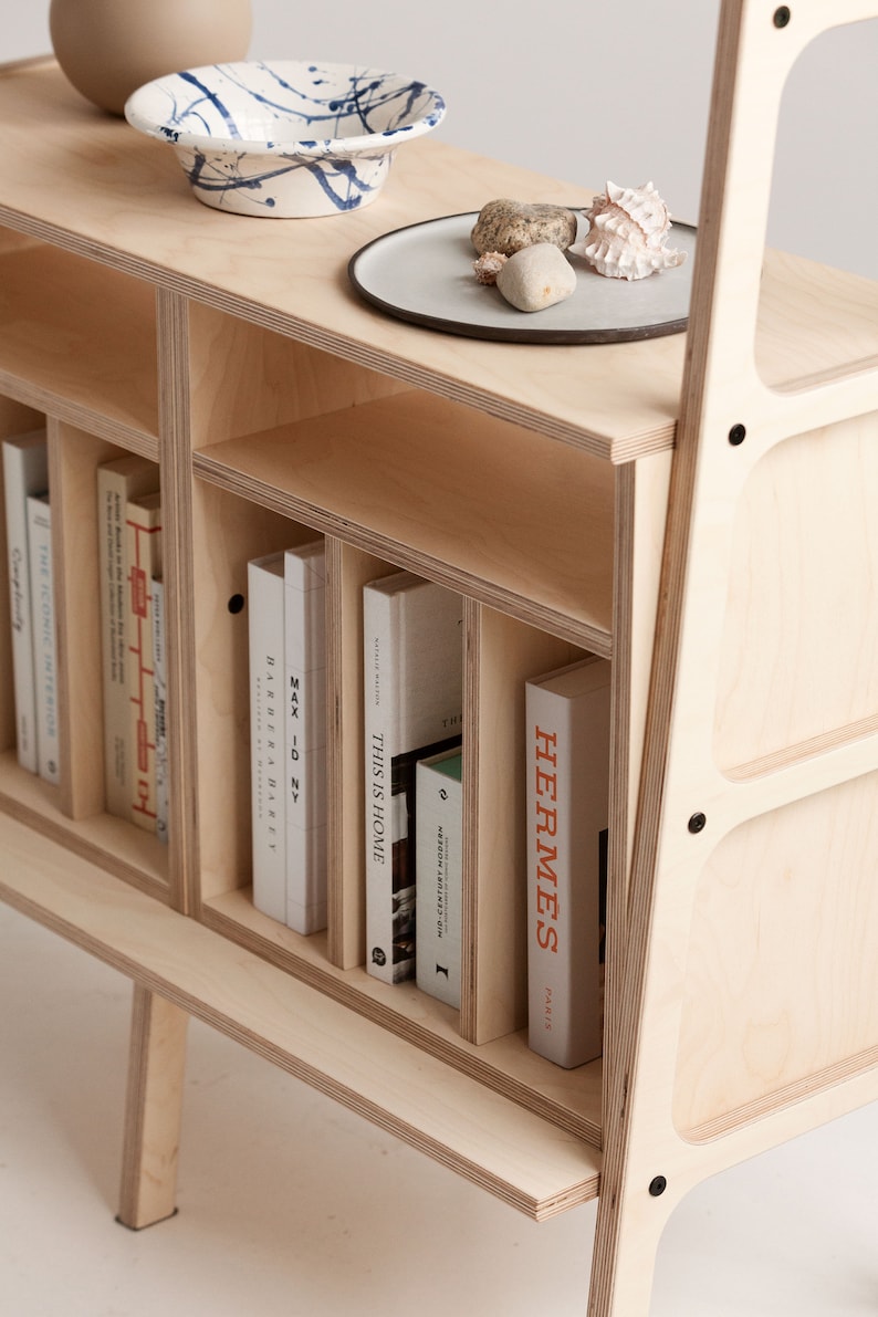 May include: A light wood bookshelf with a slanted back and a lower shelf. The bookshelf has several compartments and is filled with books, including "This is Home", "Max ID NY", "Barber & Osgerby", and "Hermes Paris".