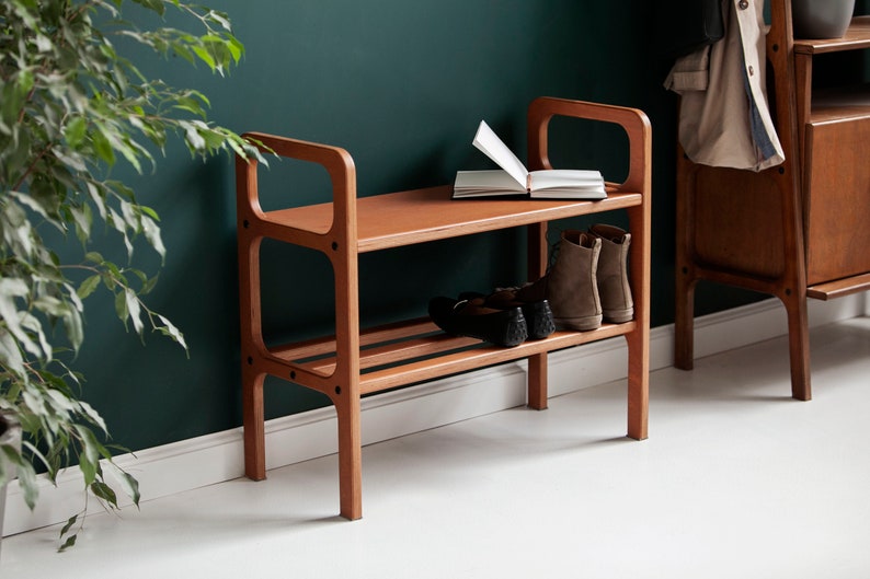 May include: A wooden shoe bench in a warm brown hue, featuring two shelves and a seat. The bench has a mid-century modern design. Brown suede boots and black shoes are placed on the shelves. An open book is on the seat.